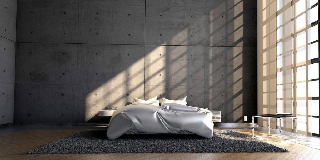 Chambre avec lit queen size drap argenté, coussins blancs, parquet, tapis gris, mur en béton, lumière naturelle