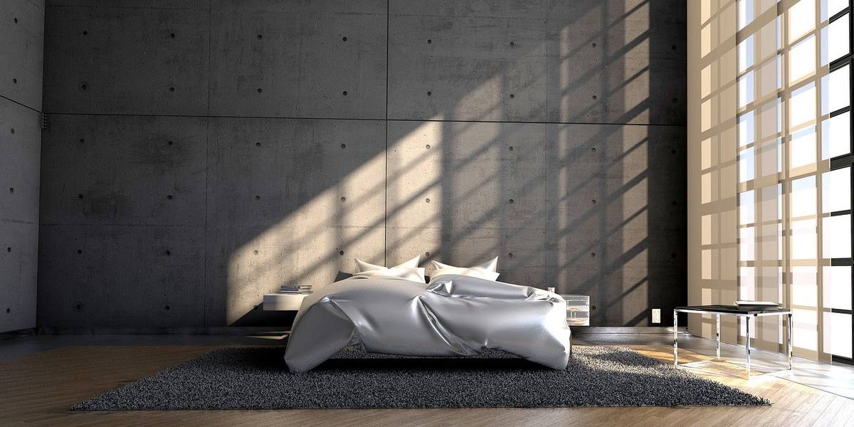 Chambre avec lit queen size drap argenté, coussins blancs, parquet, tapis gris, mur en béton, lumière naturelle
