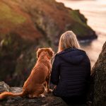 woman in black jacket sitting beside brown dog on rock near body of water during daytime